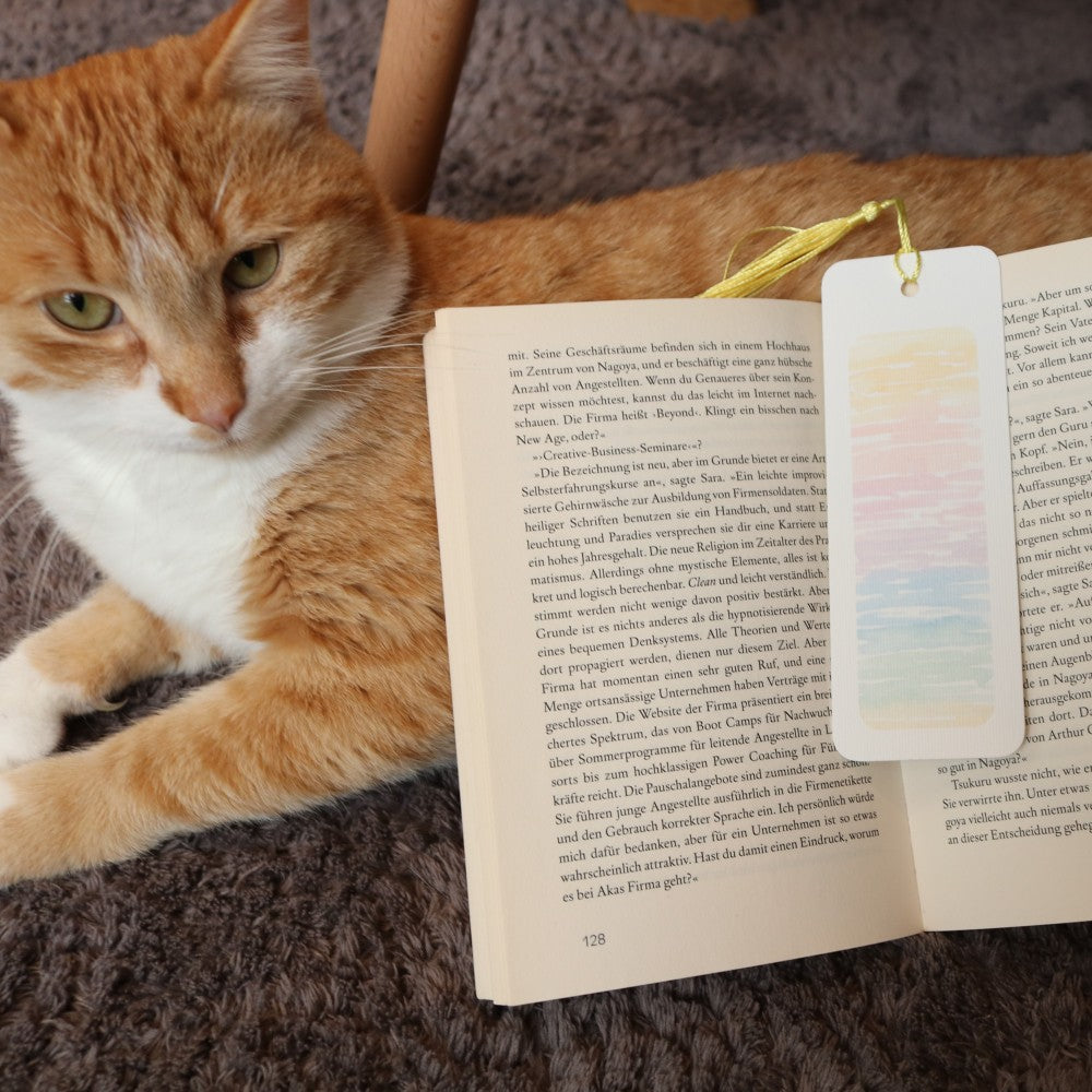 Meer-Lesezeichen mit pastelligen Aquarellfarben wie Sonnenuntergang, gelber Quaste, liegend auf einem geöffneten Buch – daneben eine entspannte rot-weiße Katze. 