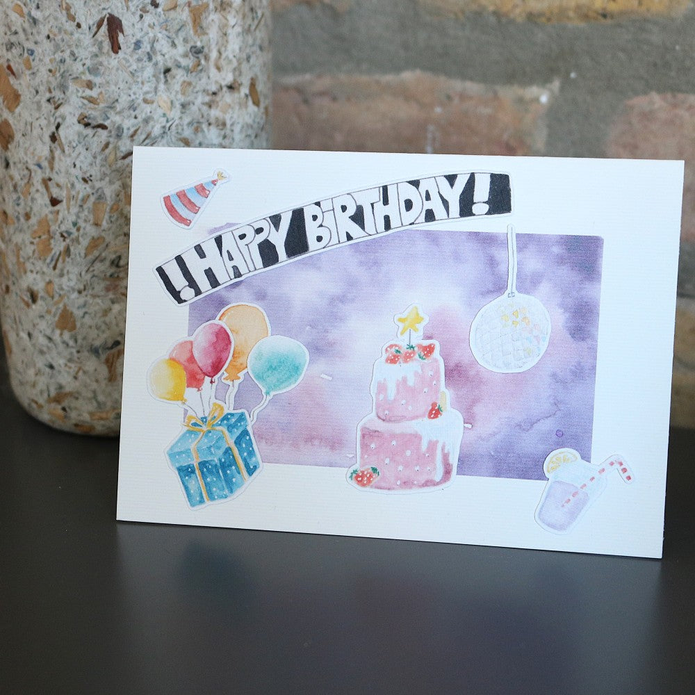 Geburtstagskarte im Querformat mit Aquarellhintergrund in Violett, darauf aufgeklebt ein pinkfarbener Kuchen, bunte Luftballons, ein Geschenk, ein Disco-Ball und ein „Happy Birthday“-Schriftzug.