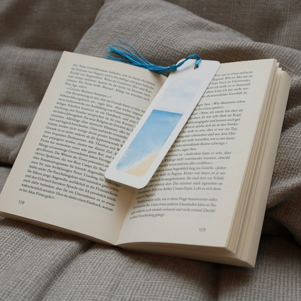 Aquarell-Lesezeichen aus dem Set Im Meer mit zarten Blau- und Sandtönen wie Himmel und Strand, mit hellblauer Quaste, liegend auf einem offenen Buch auf einer weichen Decke.