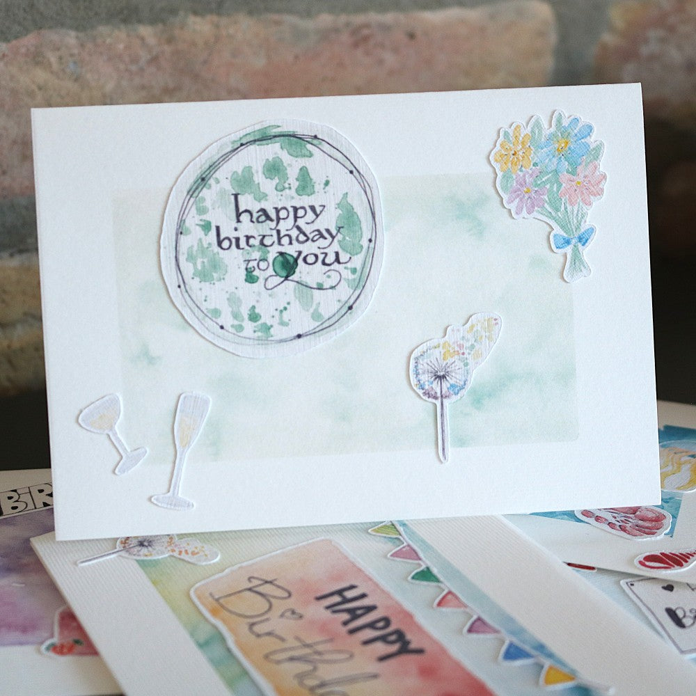 Geburtstagskarte mit zartem grünem Aquarellhintergrund, rundem „happy birthday to you“-Motiv, Blumenstrauß und kleinen Glas-Illustrationen als 3D-Elemente.