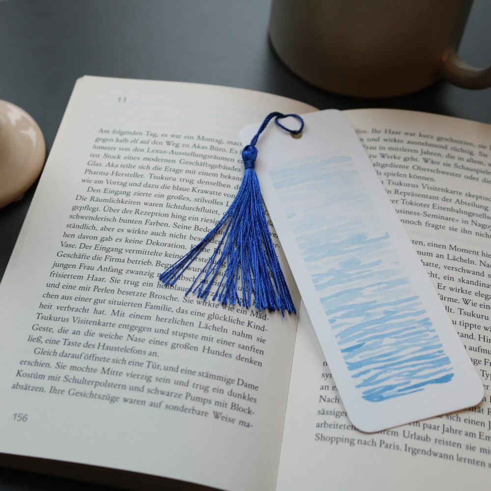 Lesezeichen-Set Im Meer: Blau gestreiftes Aquarell-Lesezeichen mit maritimer Wasseroptik und dunkelblauer Quaste auf aufgeschlagenem Buch.