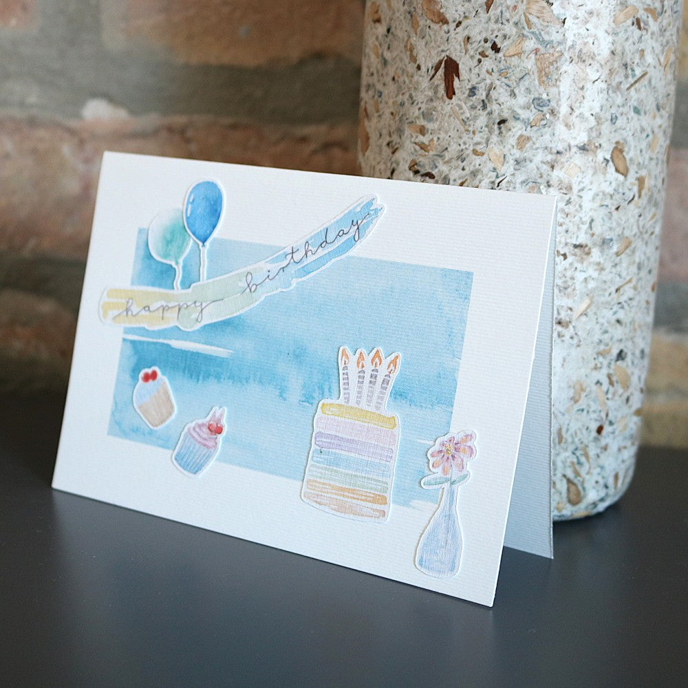 Geburtstagskarte mit blauem Aquarellhintergrund, Tortenmotiv mit Kerzen, kleinen Cupcake-Illustrationen und „Happy Birthday“-Banner als aufgeklebte 3D-Elemente.