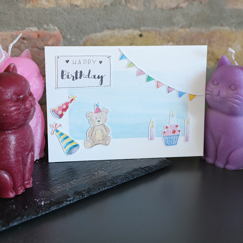 Geburtstagskarte mit hellblauem Aquarellhintergrund, Teddybär, Partyhüten, Cupcake mit Kerzen, Wimpelgirlande und „Happy Birthday“-Schriftzug als aufgeklebte 3D-Motive.