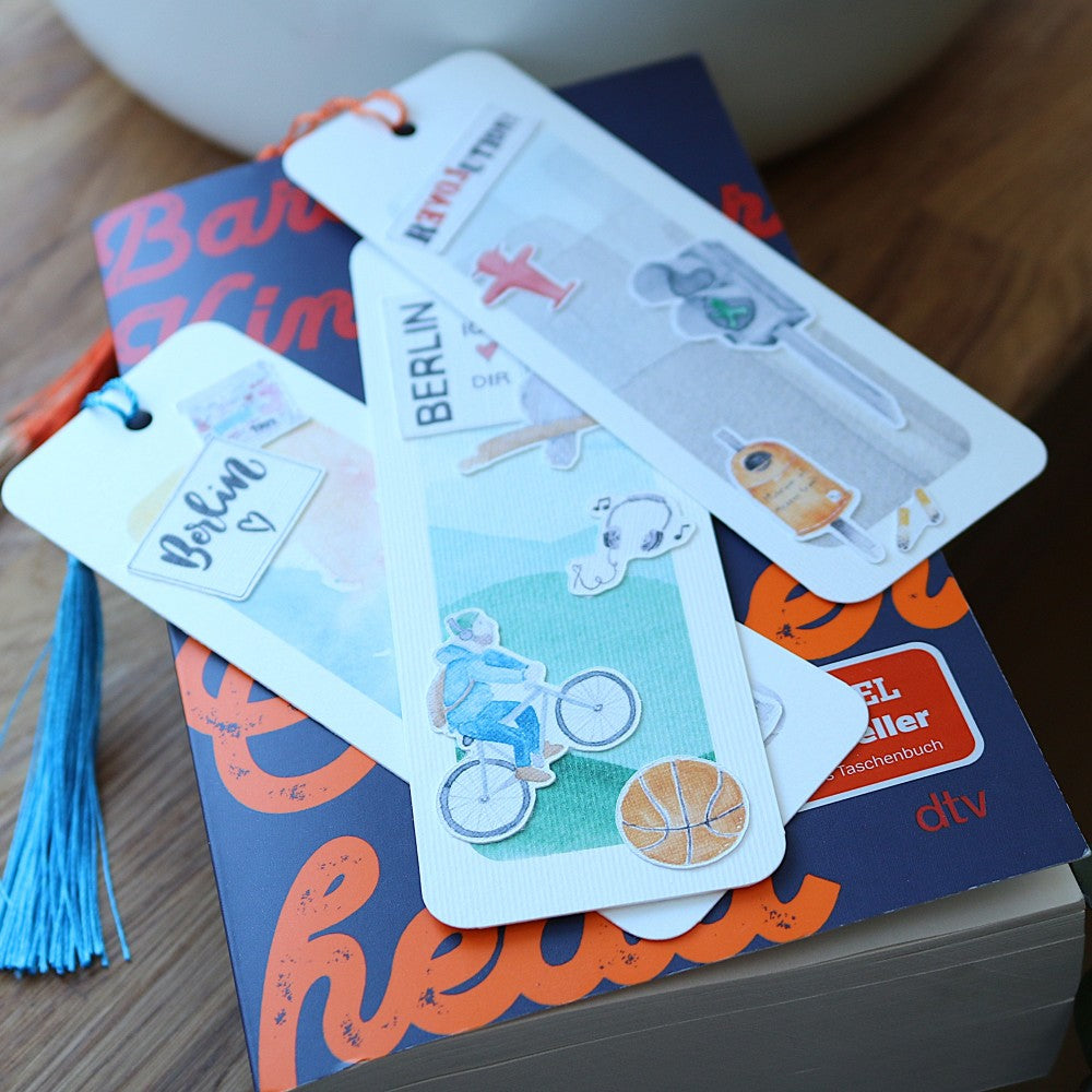 Berlin-Lesezeichen mit Aquarellmotiven wie Fahrradfahrer, Berliner Bär, Fernsehturm und Schriftzug „Berlin“, mit farbigen Quasten, auf einem Buch liegend.