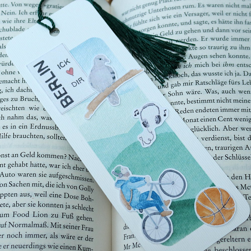 Berlin-Lesezeichen mit Aquarellillustrationen von Radfahrer, Kopfhörern, Taube und Basketball, mit grüner Quaste.