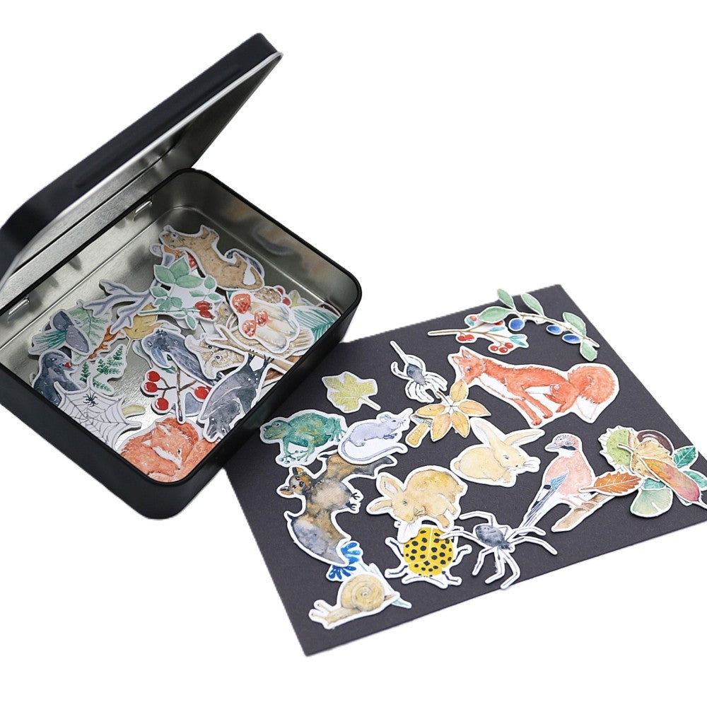 Geöffnete schwarze Metallbox der Waldbewohner Deluxe Edition mit zahlreichen Aquarell-Stickern im Inneren; weitere Motive wie Fuchs, Vögel, Hasen und Käfer und weitere Waldtiere liegen bunt gemischt auf einer dunklen Unterlage davor ausgelegt.
