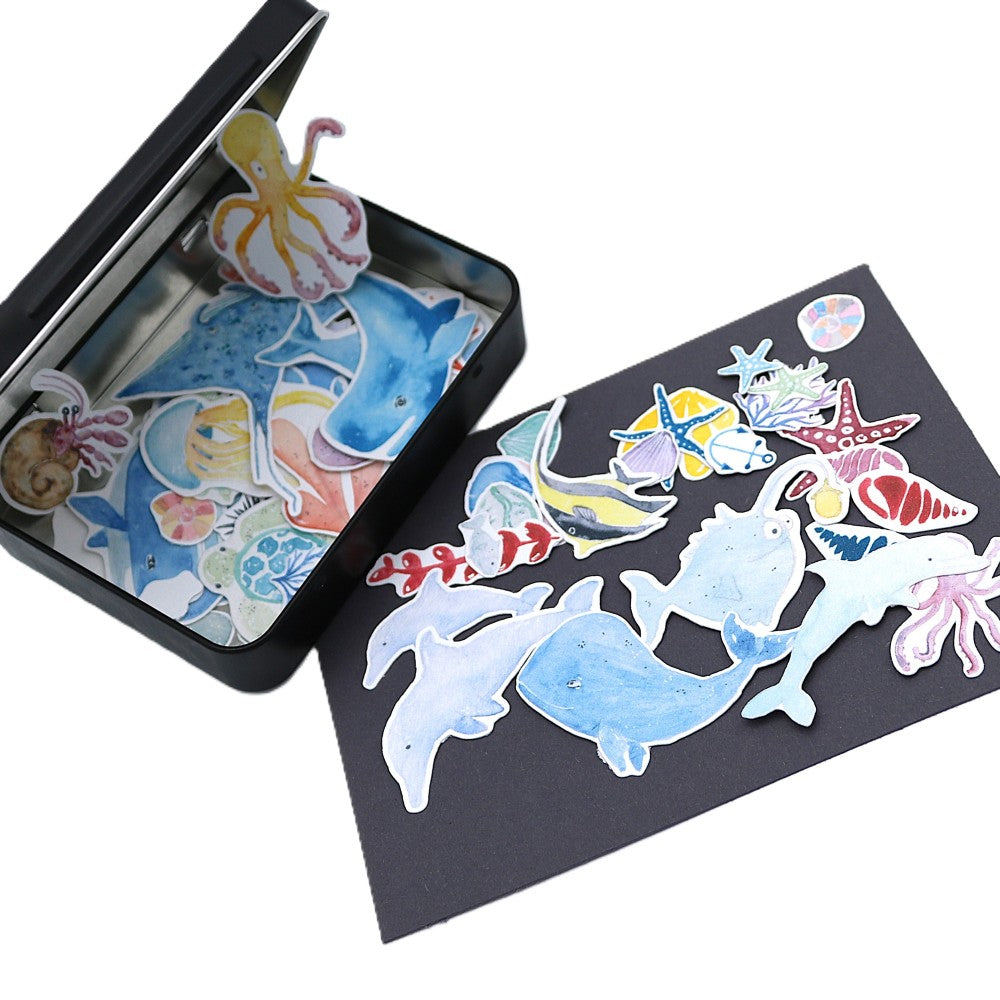 Geöffnete schwarze Metallbox der „Im Meer“ Deluxe Edition mit zahlreichen maritimen Aquarell-Stickern; im Inneren liegen bunte Unterwassermotive dicht beieinander, während vor der Box auf dunkler Unterlage größere Figuren wie Wale, Fische, Seesterne und eine Oktopus-Illustration weit aufgefächert sichtbar sind.