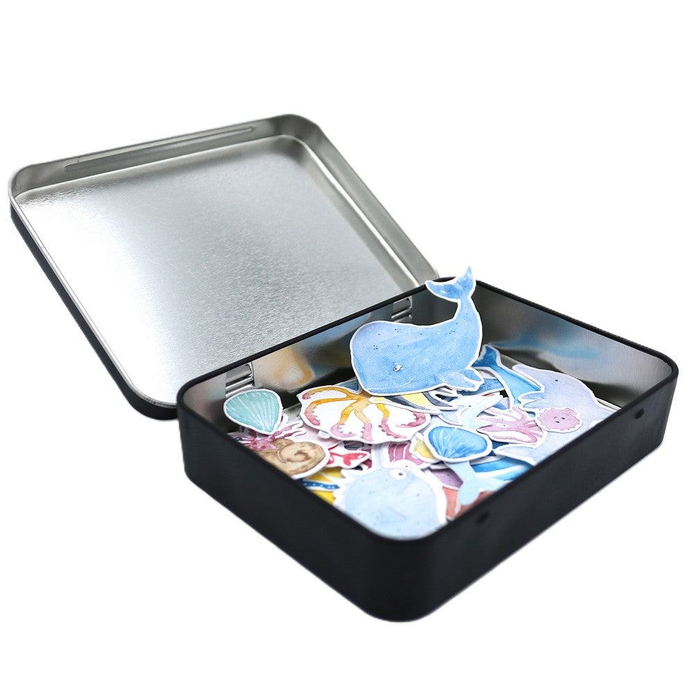 Geschlossene schwarze Metallbox der „Im Meer“ Deluxe Edition mit einem hellblauen Aquarell-Wal-Sticker auf dem Deckel; im leicht geöffneten Inneren sind farbenfrohe Meeresmotive wie Wale, Fische, Kraken und maritime Elemente als Sticker erkennbar.