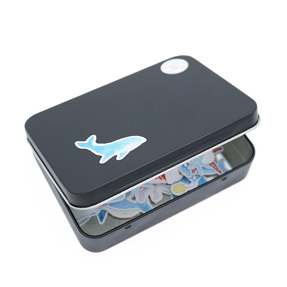 Geschlossene schwarze Metallbox der „Im Meer“ Deluxe Edition mit einem hellblauen Aquarell-Wal-Sticker auf dem Deckel; im leicht geöffneten Inneren sind farbenfrohe Meeresmotive wie Fische, Seesterne und maritime Elemente als Sticker erkennbar.