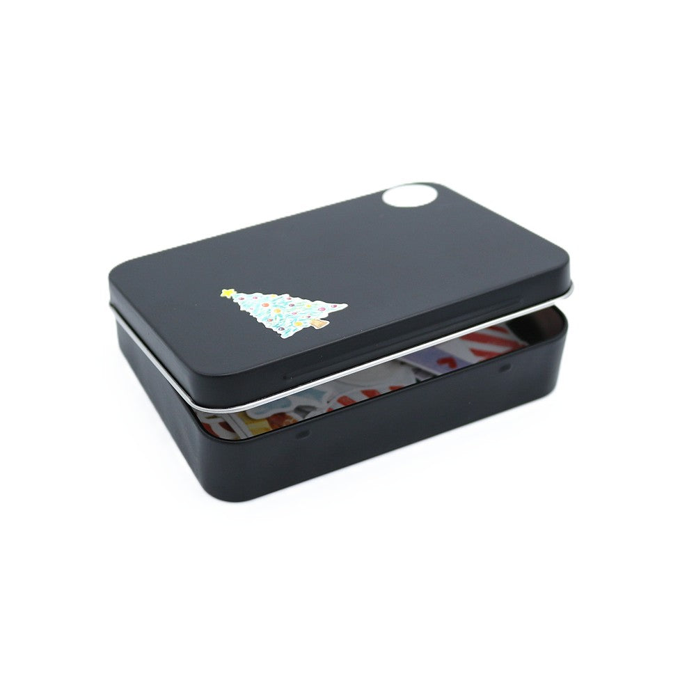 Schwarze Metallbox der Deluxe-Edition mit weihnachtlichen Stickermotiven, leicht geöffnet, sodass die farbenfrohen Aquarell-Sticker im Inneren sichtbar werden; auf dem Deckel befindet sich ein kleiner Weihnachtsbaum-Sticker.