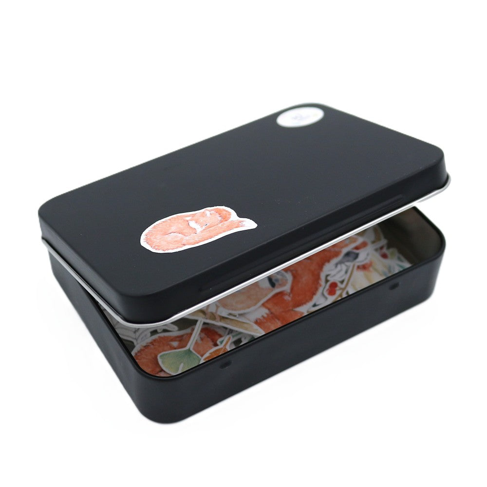 Geschlossene schwarze Metallbox der Waldbewohner Deluxe Edition, leicht geöffnet, sodass die farbenfrohen Aquarell-Sticker im Inneren sichtbar werden; auf dem Deckel klebt ein kleines Waldbewohner-Motiv.