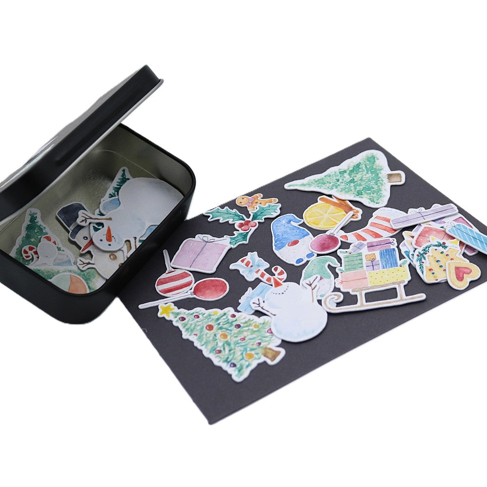 Geöffnete schwarze Metallbox der Xmas Classic Edition mit winterlichen Aquarell-Stickern wie Tannenbäumen, WIchteln, Schneemännern und Schlitten; einige Motive liegen im Inneren, weitere bunt gemischt auf einer dunklen Unterlage davor ausgelegt.