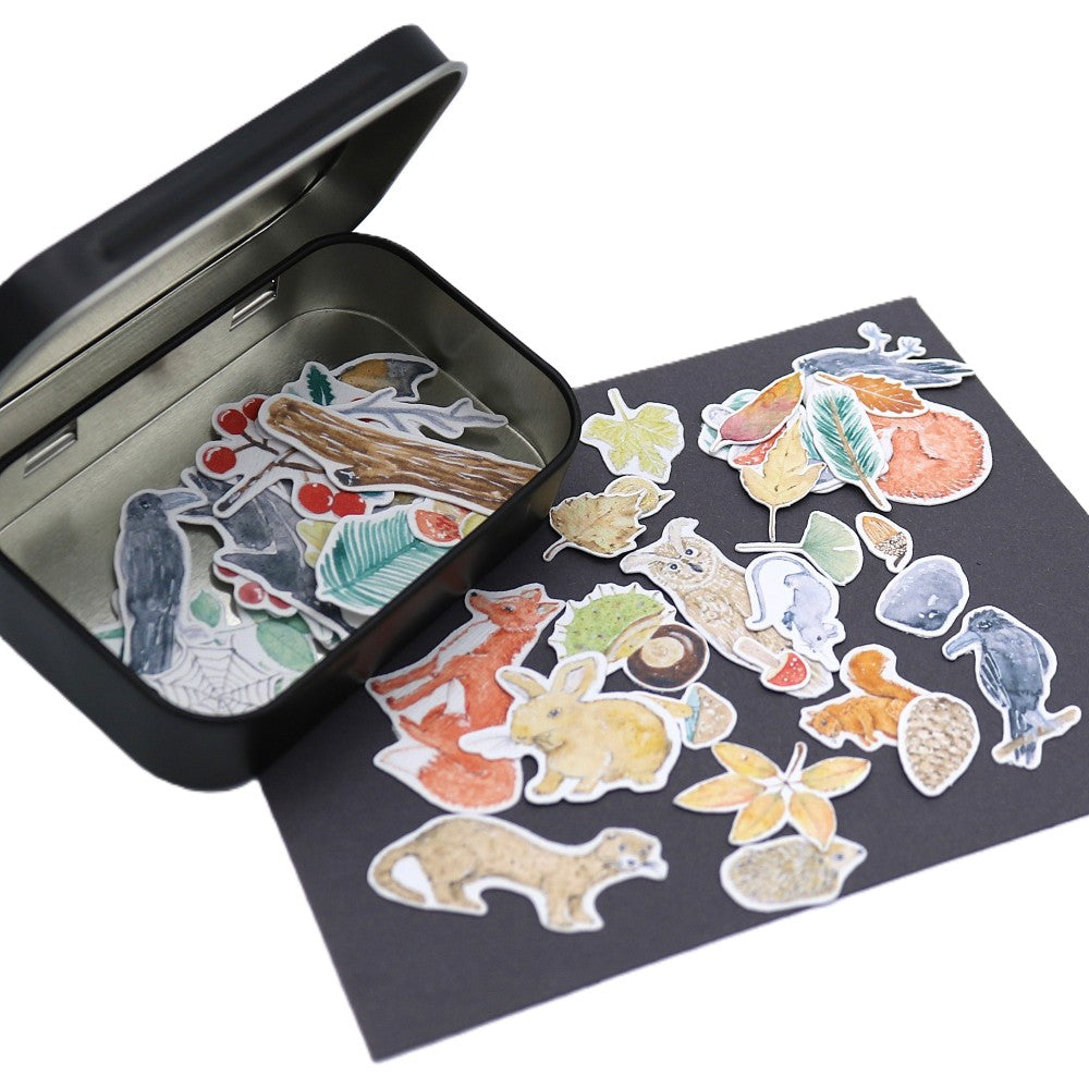 Geöffnete schwarze Metallbox der Waldbewohner Classic Edition mit zahlreichen Aquarell-Stickern im Inneren; weitere Waldbewohner-Motive wie Fuchs, Hase, Krähe, Blätter, Beeren und Pilze breit gefächert auf einer dunklen Unterlage davor ausgelegt.