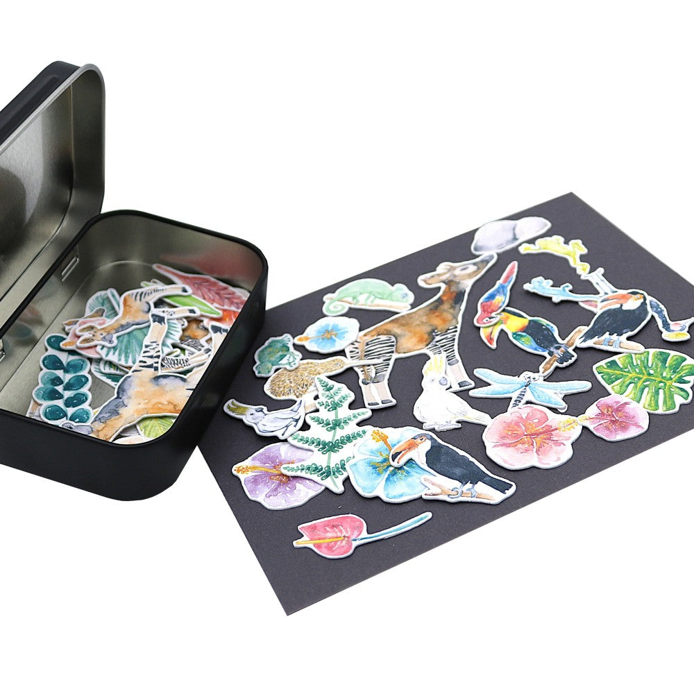 Stickerbox Tropenwelt Classic geöffnet – handillustrierte Tier- und Pflanzensticker aus Aquarellpapier ausgebreitet neben der schwarzen Metallbox.