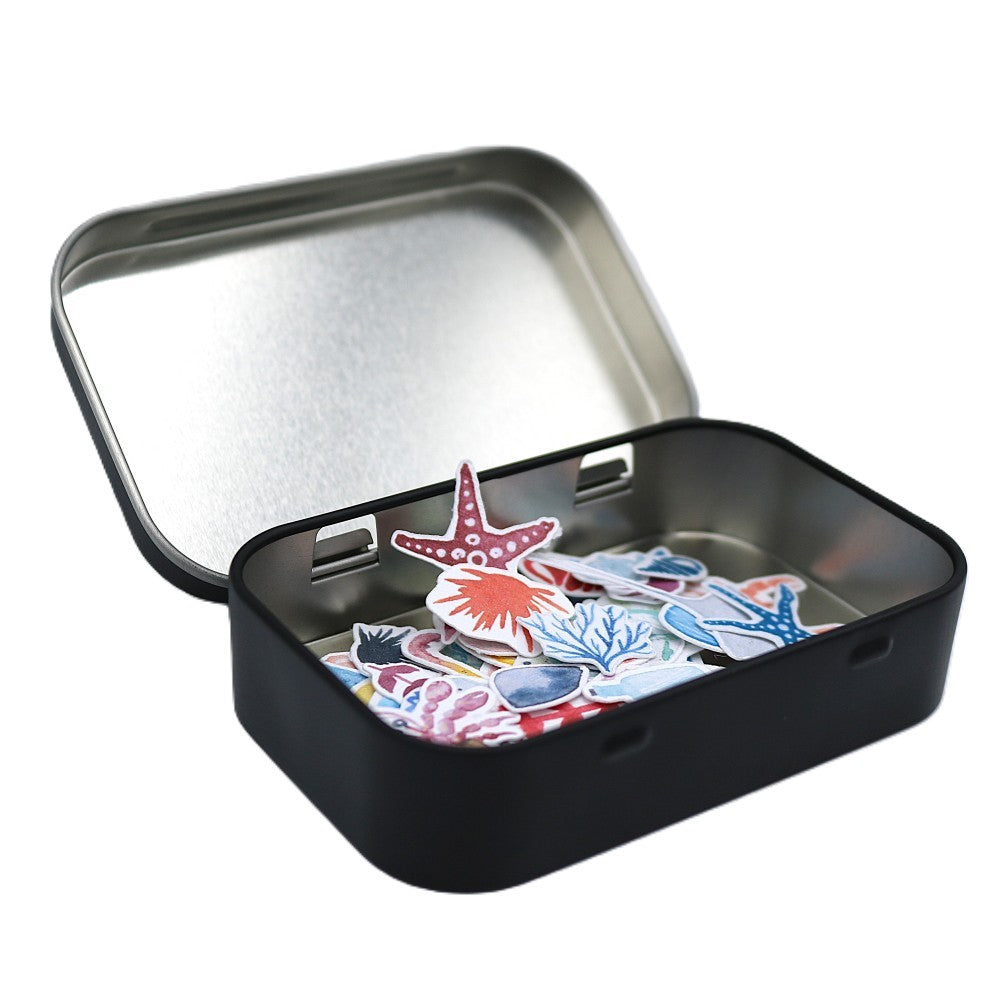 Geschlossene schwarze Metallbox der „Im Meer“ Classic Edition mit einem hellblauen Aquarell-Wal-Sticker auf dem Deckel; im leicht geöffneten Inneren sind farbenfrohe Meeresmotive wie Wale, Fische, Seesterne und maritime Elemente als Sticker erkennbar. 