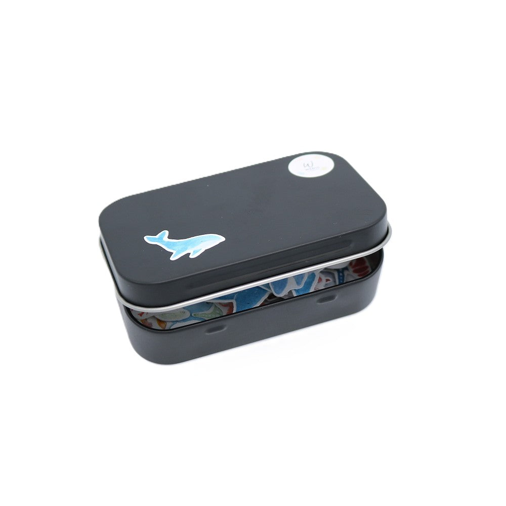 Geschlossene schwarze Metallbox der „Im Meer“ Classic Edition mit einem hellblauen Aquarell-Wal-Sticker auf dem Deckel; im leicht geöffneten Inneren sind farbenfrohe Meeresmotive wie Fische, Seesterne und maritime Elemente als Sticker erkennbar.