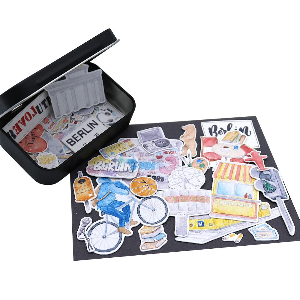 Offene schwarze Metallbox mit selbstklebenden Mini-Aquarell-Stickern mit Berlin-Motiven, ausgebreitet auf dunklem Untergrund