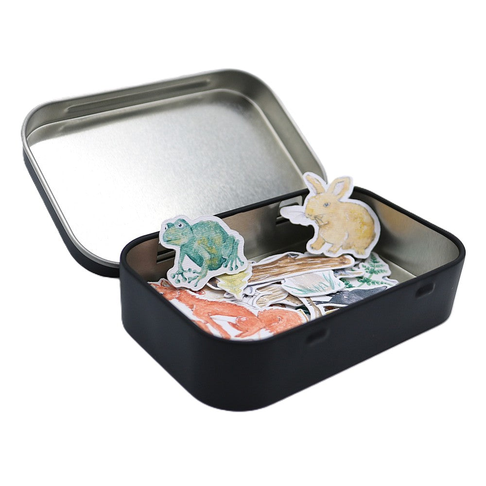 Geöffnete schwarze Metallbox der Waldbewohner Classic Edition mit sichtbaren Aquarell-Stickern wie Frosch, Hase, Fuchs, Blättern und weiteren Waldbewohner-Motiven; einige Sticker stehen aufrecht am Rand der Box, weitere liegen locker darin.