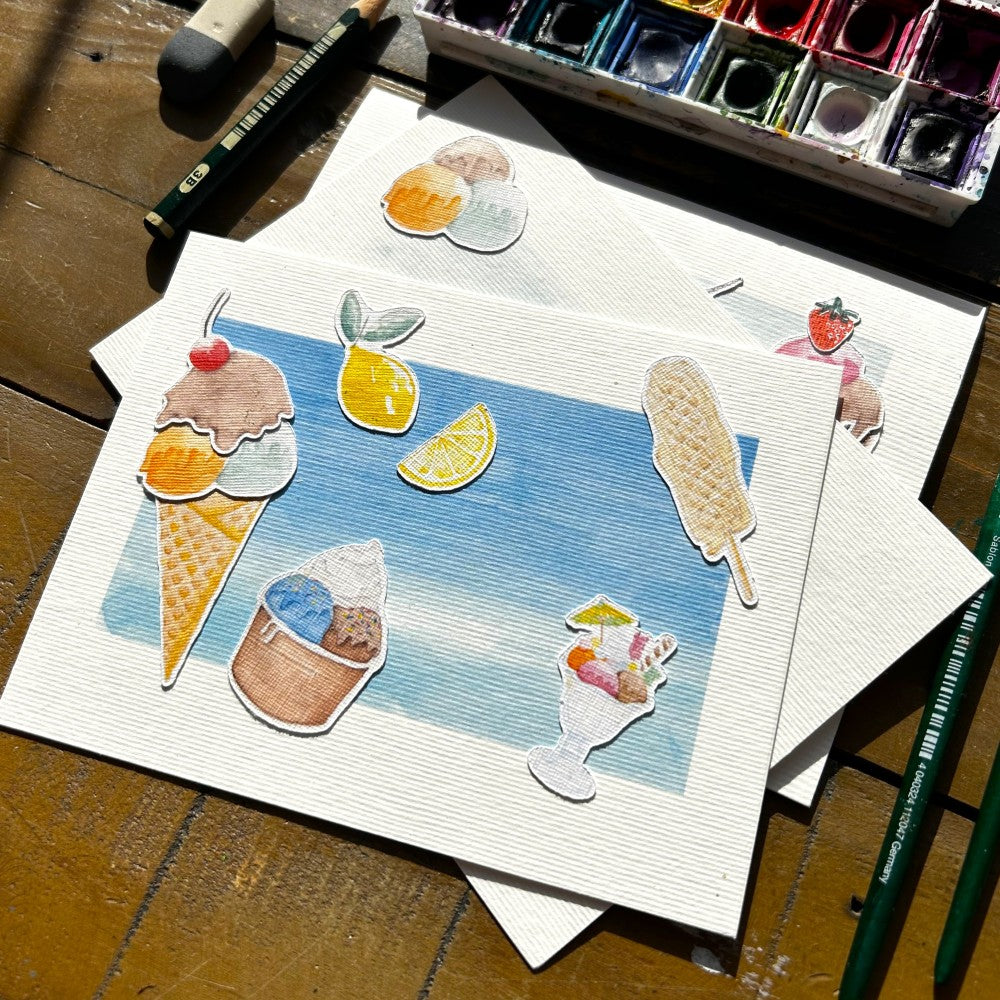 Aquarellkarte mit Eismotiven, wie Eisbecher und Eiswaffel auf einem Tisch liegend, dahinter ein Aquarell Farbkasten, Bleistift und Radiergummi 