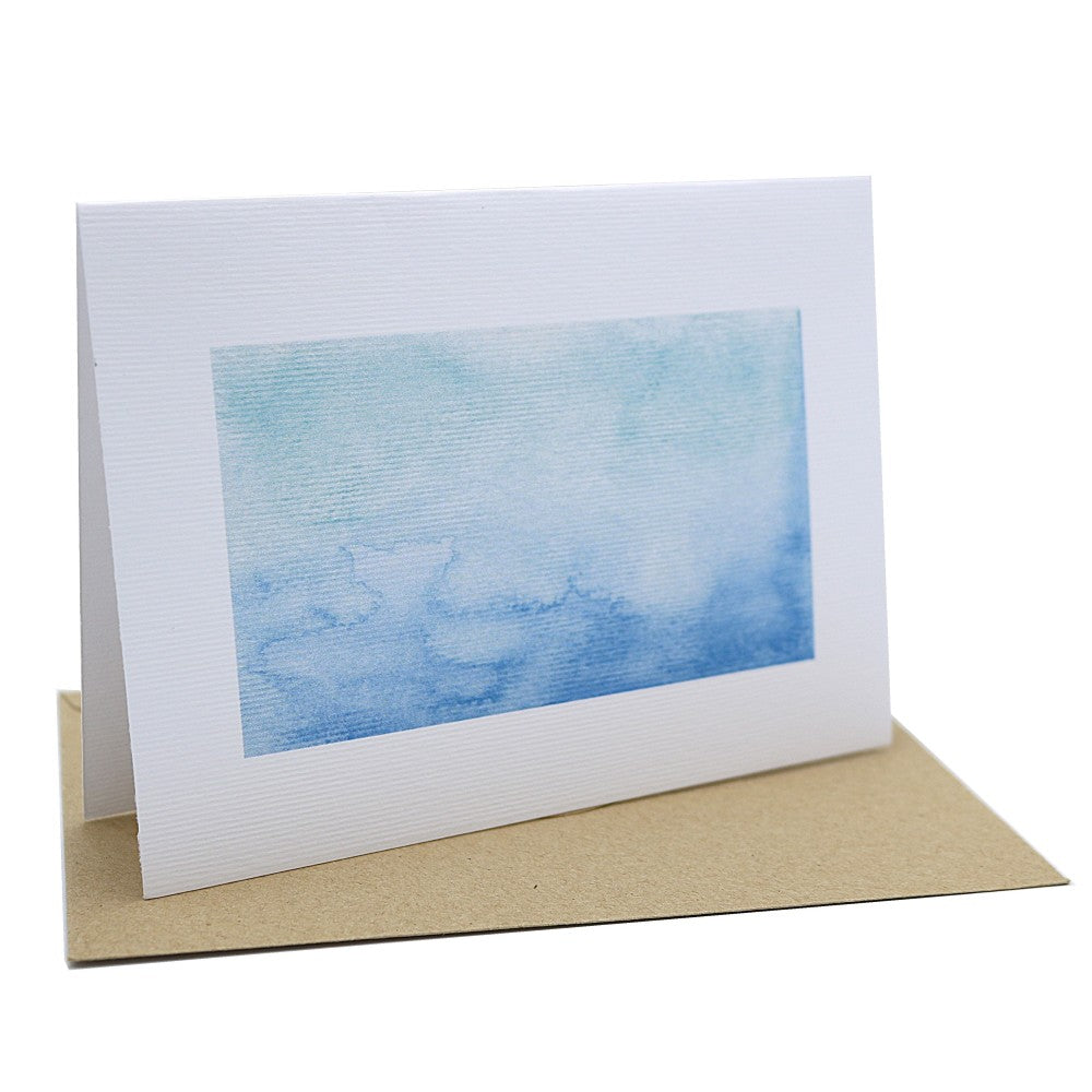 Aufgestellte Klappkarte aus strukturiertem Aquarellpapier mit blauem Meeresverlauf, dazu ein liegender Umschlag aus Kraftpapier.