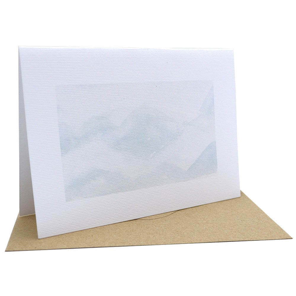 Aufgestellte Klappkarte mit winterlichem Aquarellhintergrund auf strukturiertem Aquarellpapier. Zu sehen ist eine zarte winterliche Bergandschaft in zartem grau und blau. Darunter liegt ein passender Kraftpapier-Umschlag. Der Hintergrund ist ein Detailmotiv aus dem Weihnachtskarten-Set und kommt ohne zusätzliche Motive.