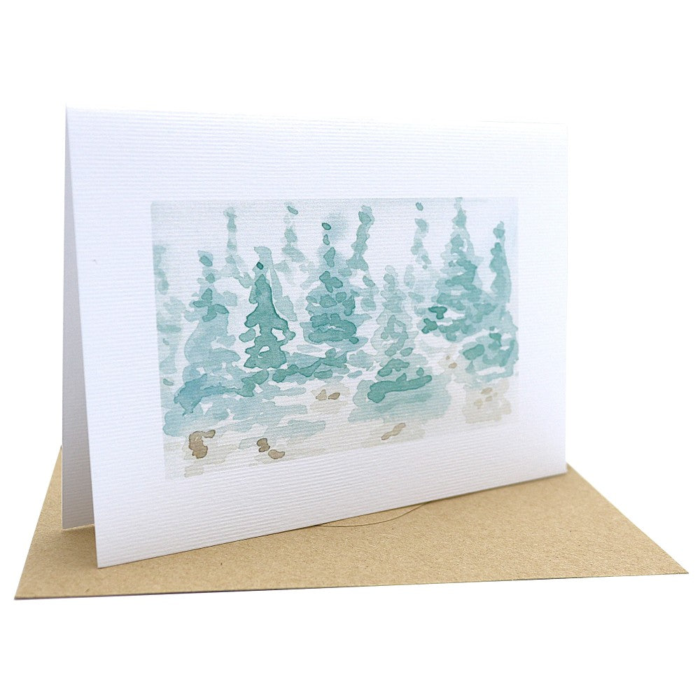 Klappkarte aufgestellt mit winterlichem Aquarellhintergrund auf strukturiertem Aquarellpapier. Zu sehen ist eine zarte Waldszene aus angedeuteten Tannen in kühlen Grüntönen. Darunter liegt ein passender Kraftpapier-Umschlag. Der Hintergrund ist ein Detailmotiv aus dem Weihnachtskarten-Set und kommt ohne zusätzliche Motive.