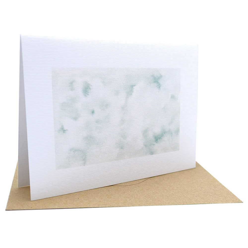 Klappkarte aus Aquarellpapier mit winterlichem Aquarellhintergrund in zarten Grün- und Weißtönen; im Vordergrund liegt ein Kraftpapier-Umschlag auf weißem Hintergrund.