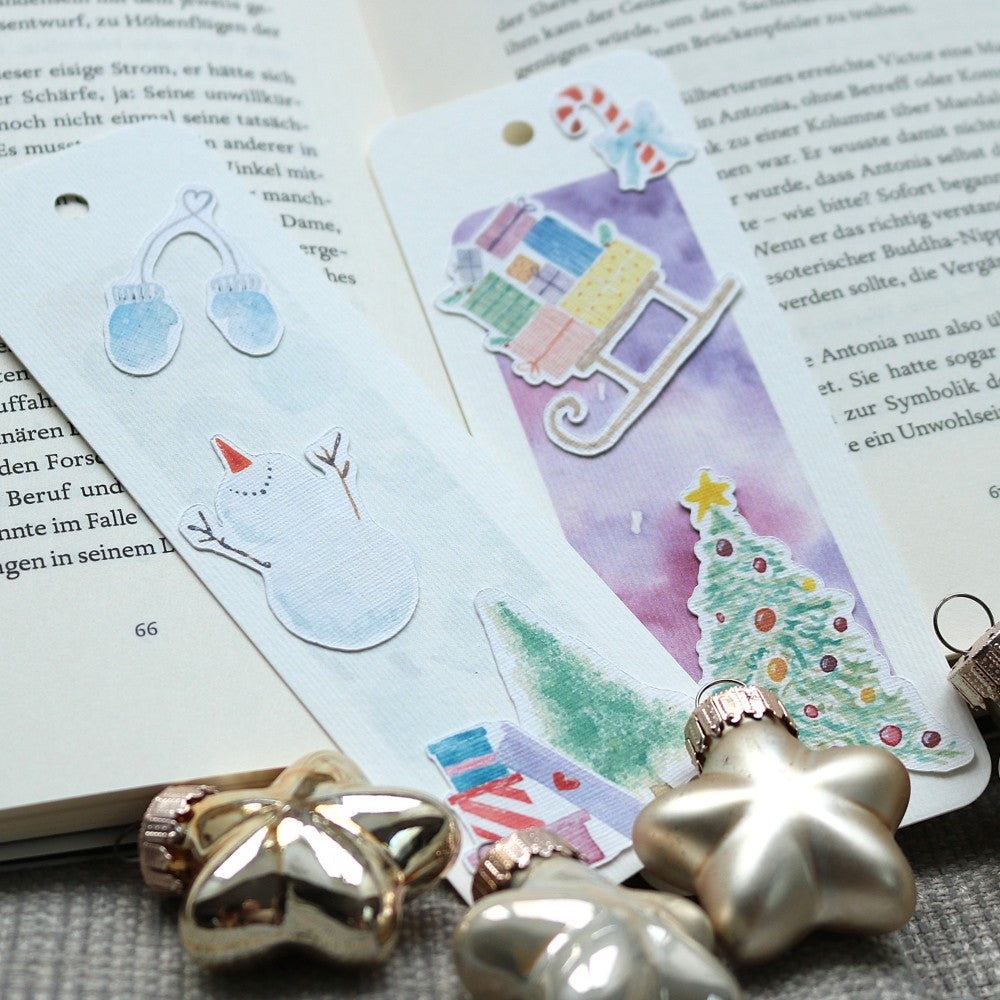 Zwei weihnachtliche Lesezeichen aus Aquarellpapier auf einem geöffneten Buch; Motive wie Schneemann, Fäustlinge, Geschenkeschlitten und geschmückter Weihnachtsbaum; dekoriert mit kleinen goldenen Stern-Ornamenten davor.