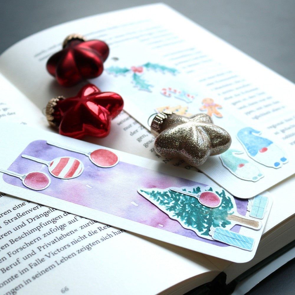 Zwei weihnachtliche Lesezeichen aus Aquarellpapier auf einem offenen Buch, dekoriert mit roten Stern-Christbaumkugeln; Motive wie Zuckerstangen und geschmückter Tannenbaum sichtbar.