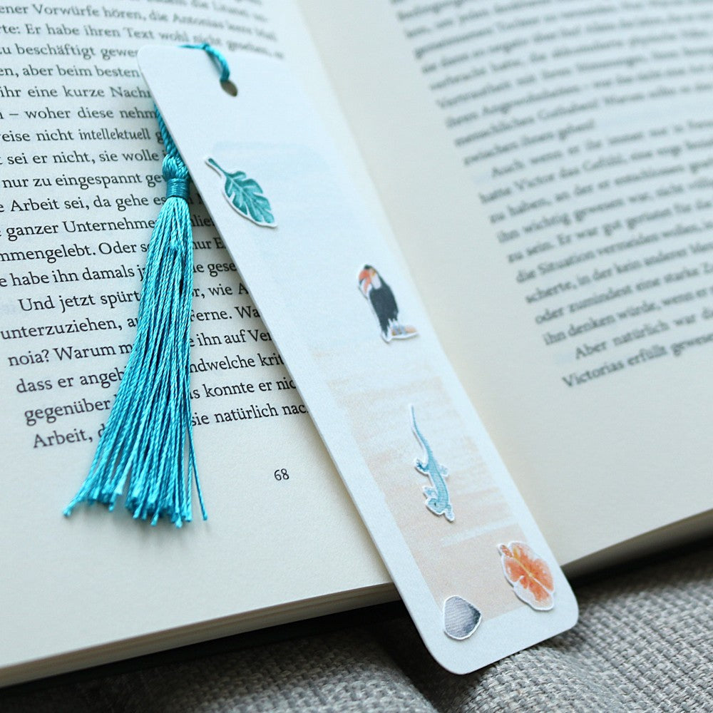 Aquarell-Lesezeichen Tropenwelt mit blauer Quaste und tropischen Stickermotiven – Blatt, Vogel, Eidechse, Blüte – liegend auf einem geöffneten Buch