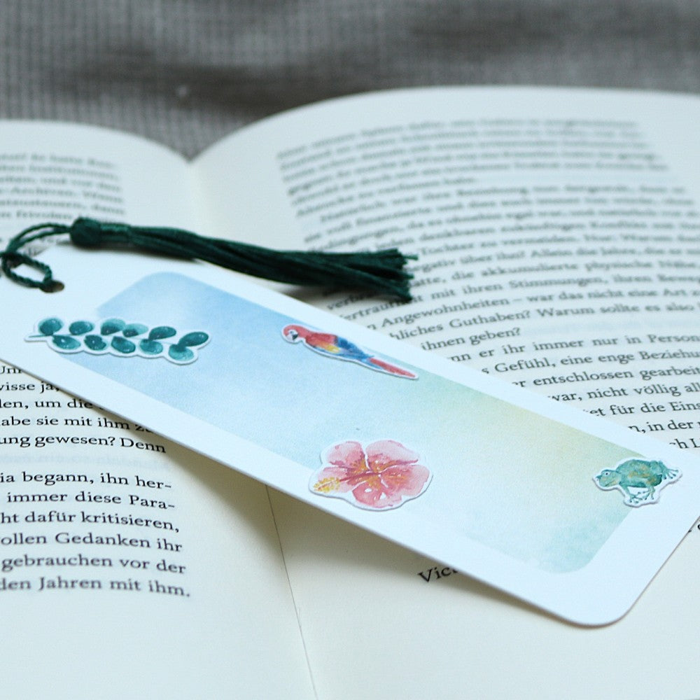 Aquarell-Lesezeichen Tropenwelt mit grüner Quaste und tropischen Stickermotiven – Papagei, Blüte, Eukalyptus und Chamäleon – auf einem geöffneten Buch
