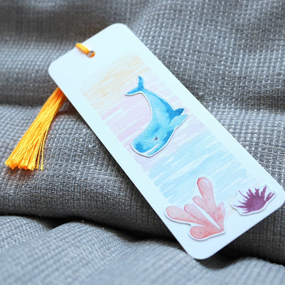 Lesezeichen Im Meer: Aquarell-Lesezeichen mit blauem Wal und Korallenmotiven auf pastellfarbenem Hintergrund, mit gelber Quaste
