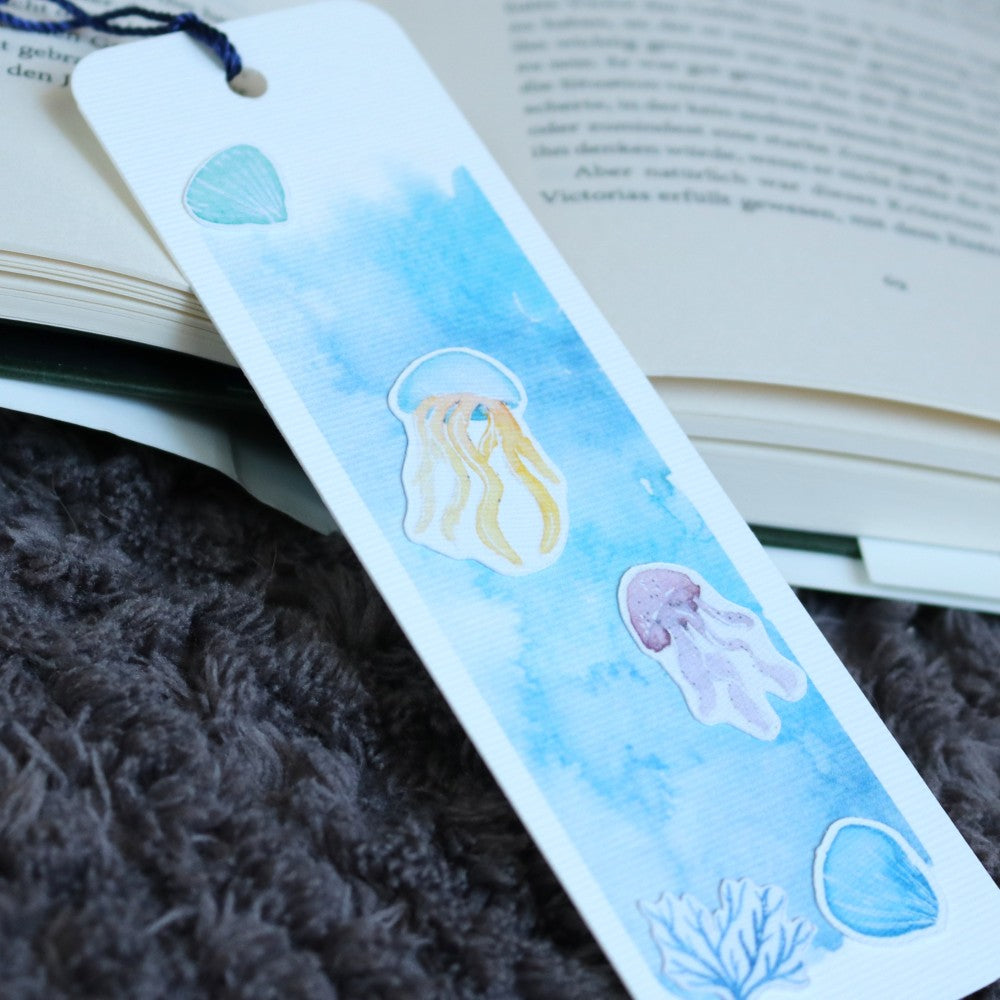 Lesezeichen Im Meer: Blaues Aquarell-Lesezeichen mit zwei Quallen und Meeresmotiven wie Muschel und Koralle, auf einem geöffneten Buch liegend