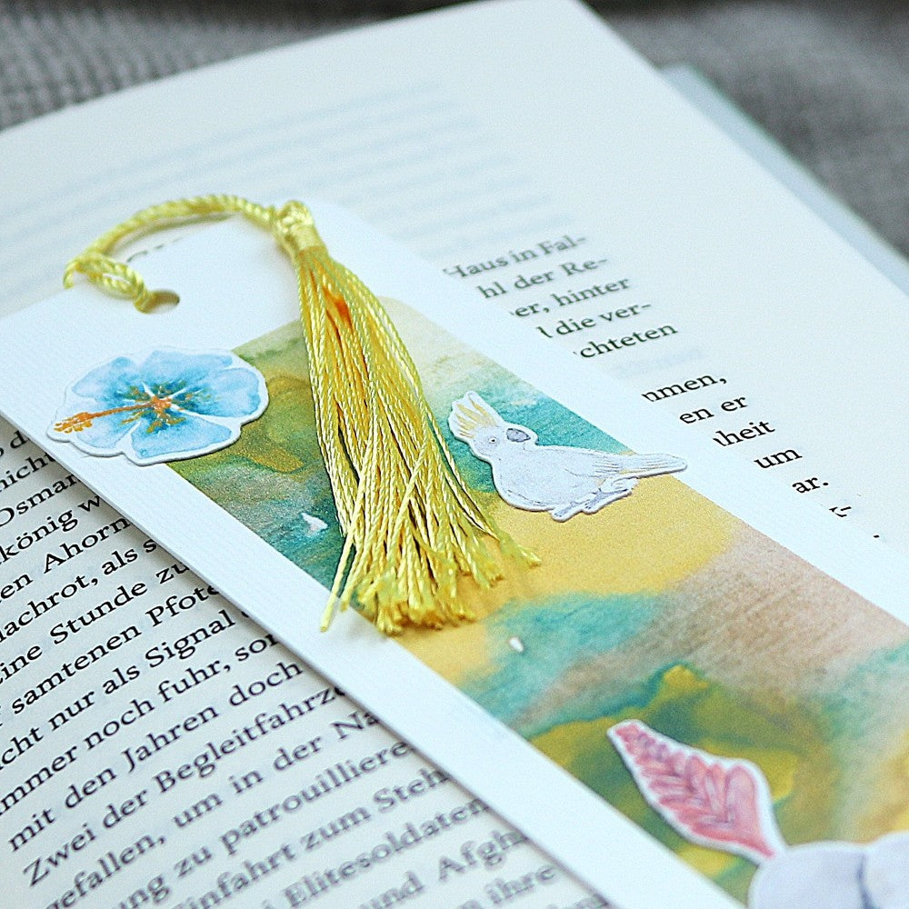 Aquarell-Lesezeichen Tropenwelt mit goldfarbener Quaste, Vogel- und Blütenstickern – auf einem geöffneten Buch liegend, Detailaufnahme