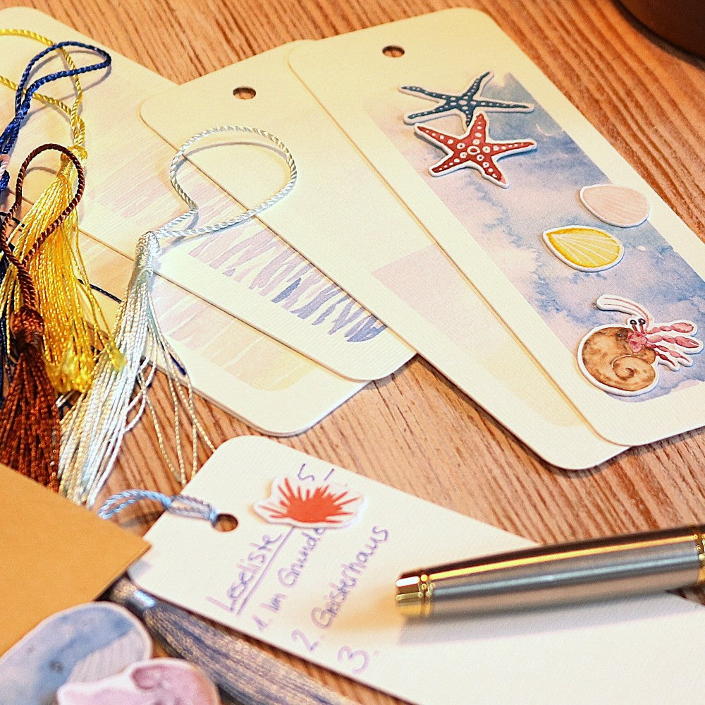 mehrere Lesezeichen liegend, mit Aquarellhintergründen und Meeresmotiven, eines davon umgedreht und beschrieben, daneben ein Stift sowie passende Lesezeichenbändchen mit Quaste.