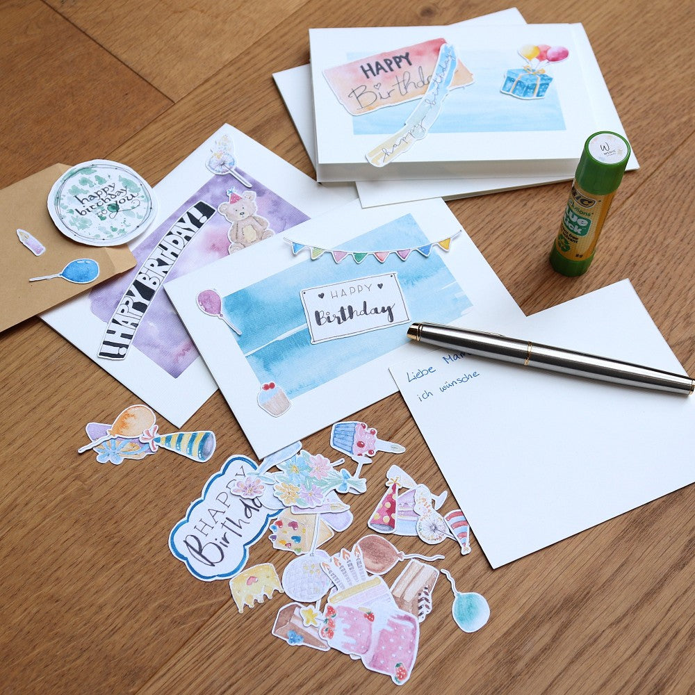 Geburtstags-Kartenset mit gestalteter Karte, Aquarell-Stickern, Klebestift und Umschlag auf Tisch arrangiert
