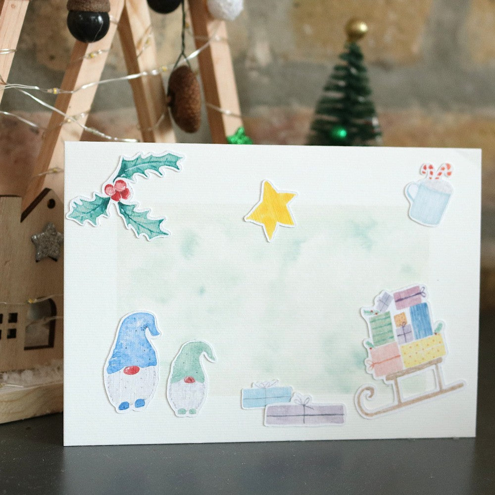 Klappkarte mit winterlichem Aquarellhintergrund und ausgeschnittenen Weihnachtsmotiven wie Stechpalme, Stern, Geschenken, Schlitten und zwei Wichteln; Deko im Hintergrund.