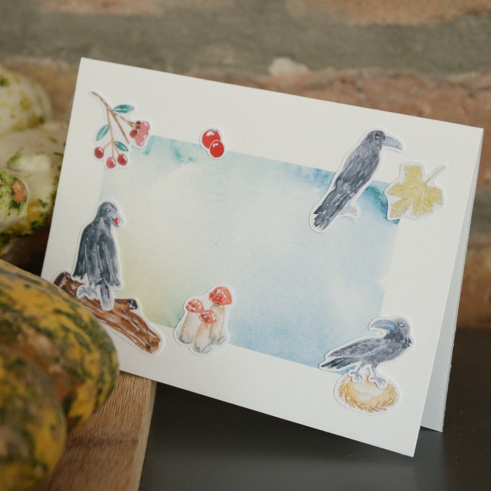 Klappkarte mit hellblau-grünem Aquarell-Hintergrund, dekoriert mit ausgeschnittenen Motiven aus dem Waldbewohner-Set: zwei Krähen, Pilze, Blätter und rote Beeren; die Karte steht vor einer rustikalen Steinwand und liegt teilweise auf einem Kürbis
