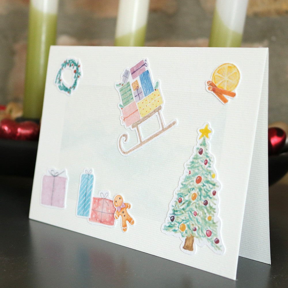 Klappkarte mit winterlichem Aquarellhintergrund und ausgeschnittenen Weihnachtsmotiven wie Schlitten, Geschenken, Kranz, Zimt-Orangenscheibe, Lebkuchenmann und geschmücktem Tannenbaum; stimmungsvolle Deko im Hintergrund
