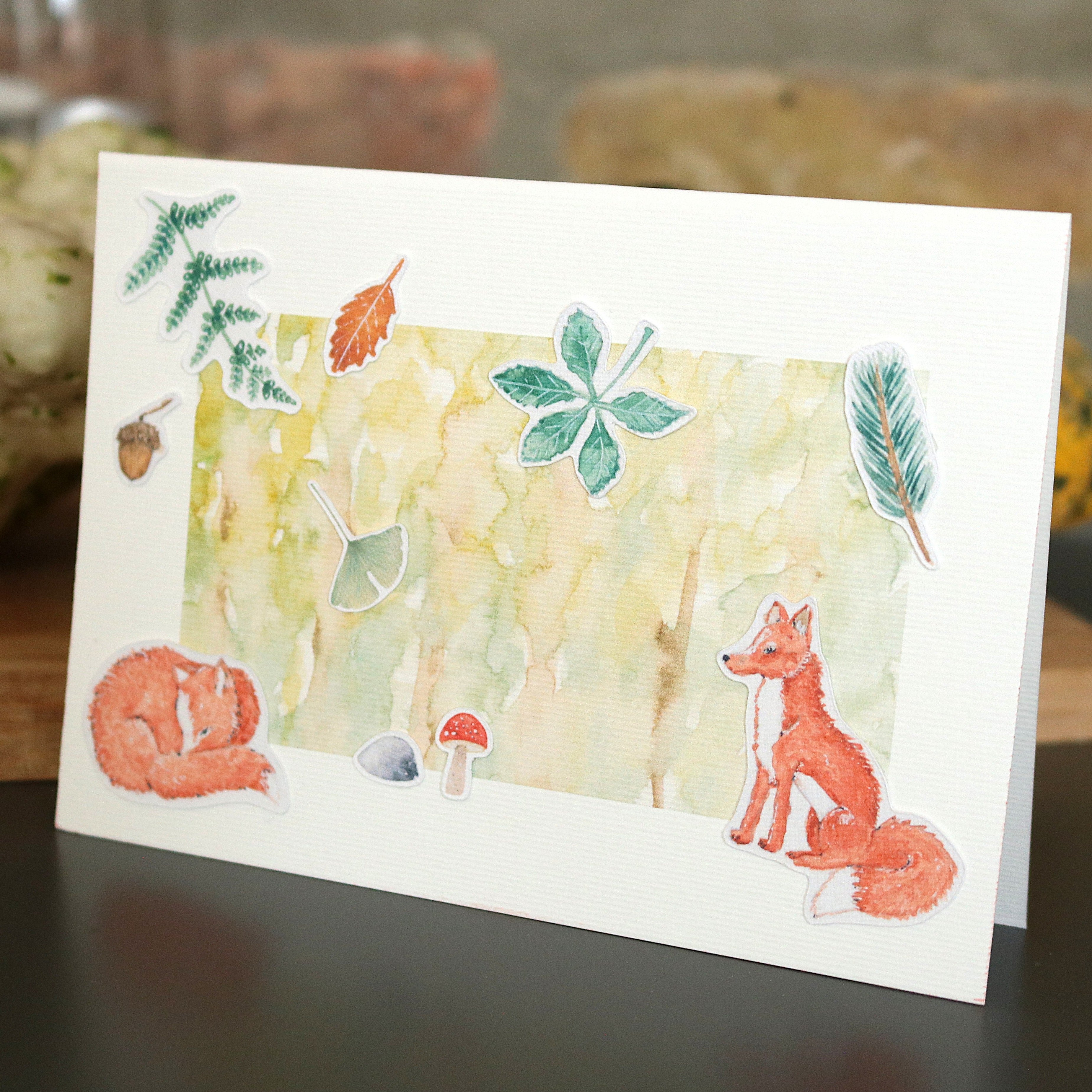 Klappkarte mit Wald-Aquarellhintergrund, verziert mit Motiven aus dem Set „Waldbewohner“ – Fuchs, schlafendes Eichhörnchen, Blätter, Feder und kleine Waldteile – Karte steht auf einem Tisch vor unscharfem Hintergrund