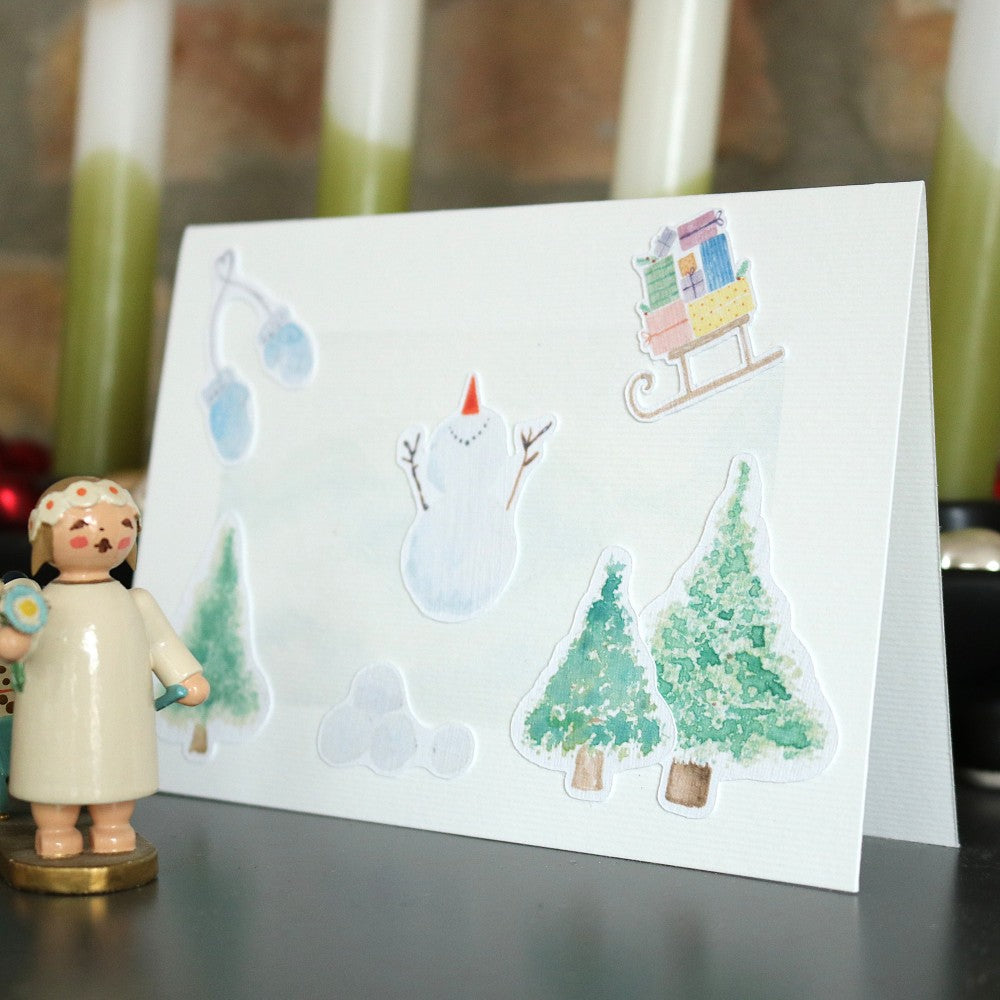 Klappkarte mit winterlichem Aquarellhintergrund und ausgeschnittenen Motiven wie Schneemann, Tannenbäumen, Schlitten und Handschuhen; Dekofigur links daneben