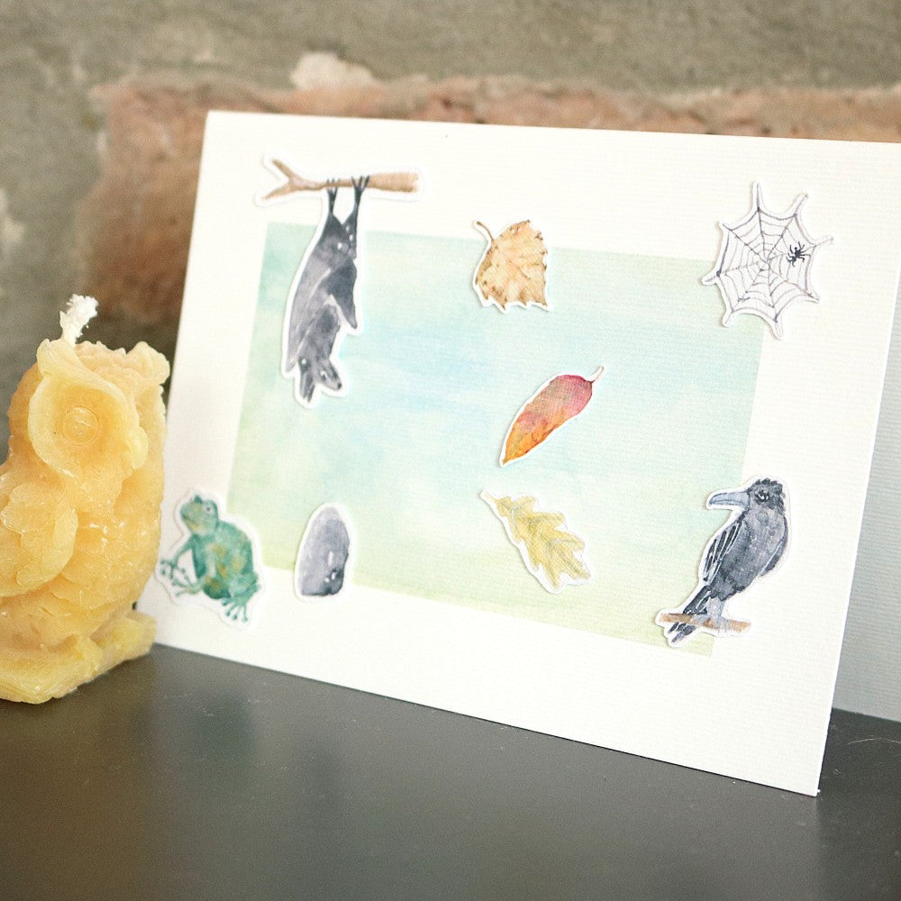 Klappkarte mit Wald-Aquarellhintergrund, dekoriert mit einzelnen Motivelementen zum Aufkleben aus dem Set „Waldbewohner“, Karte steht auf einem Tisch vor Deko