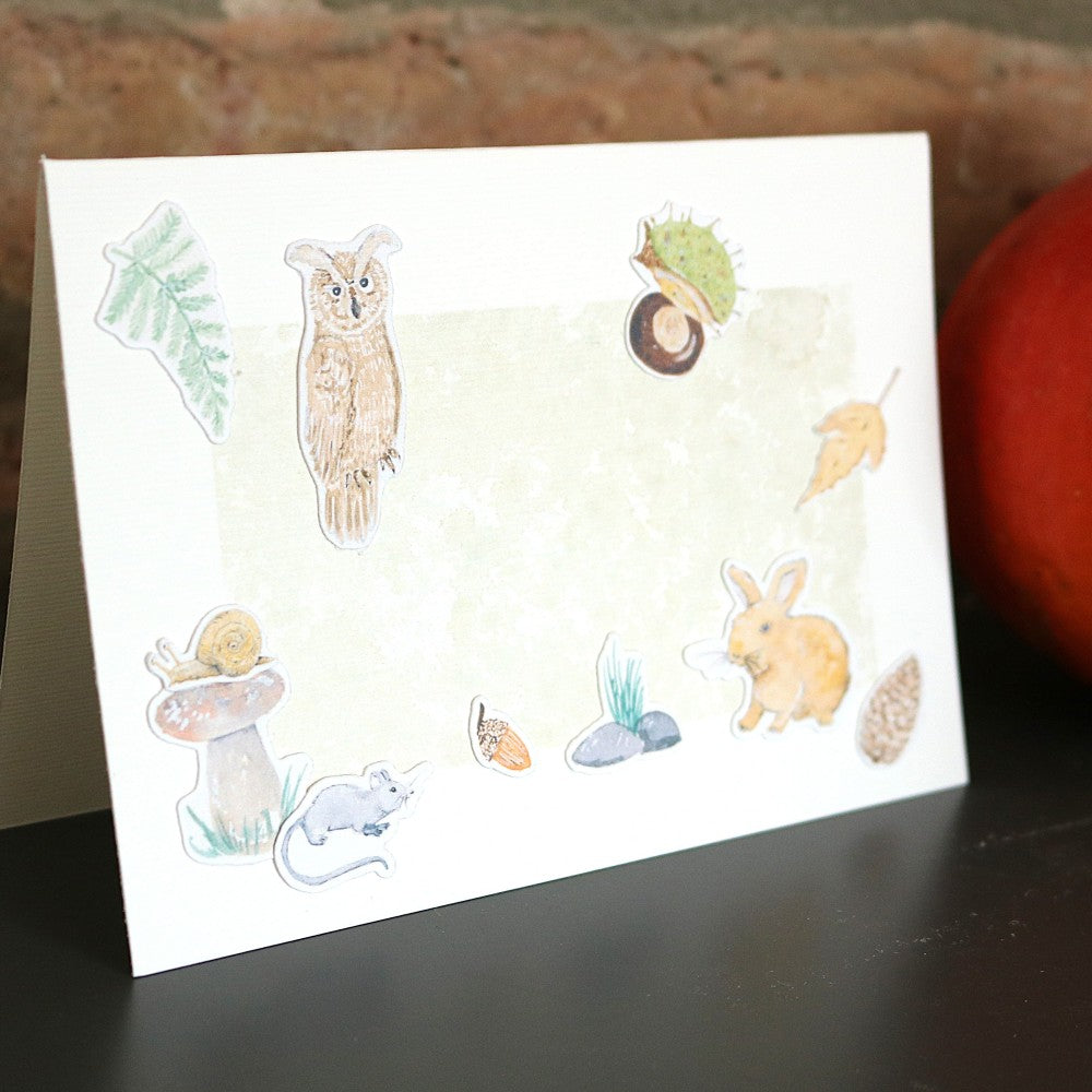 Klappkarte mit Wald-Aquarellhintergrund, verziert mit ausgeschnittenen Motiven aus dem Set „Waldbewohner“ – Eule, Hase, Maus, Pilz, Schnecke, Blätter und Kastanienhülle; Karte steht auf dunkler Oberfläche vor Ziegelwand und Kürbisdeko