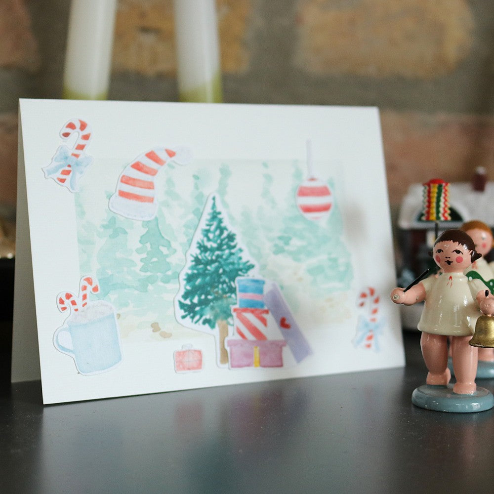Klappkarte mit winterlichem Aquarell-Hintergrund und ausgeschnittenen Weihnachtsmotiven wie Tannenbaum, Zuckerstangen, Mütze, Geschenken und Christbaumkugeln; rechts daneben stehen kleine Holzfiguren als Deko, die nicht zum Produkt gehören