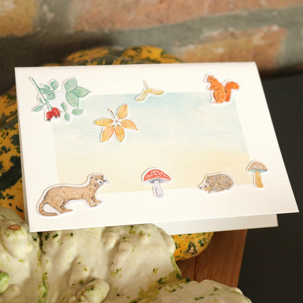Klappkarte mit Wald-Aquarellhintergrund, dekoriert mit ausgeschnittenen Motiven aus dem Set „Waldbewohner“ – darunter Igel, Marder, Pilze, Blätter und ein rotes Eichhörnchen, Karte steht auf Kürbissen vor Ziegelwand-Hintergrund