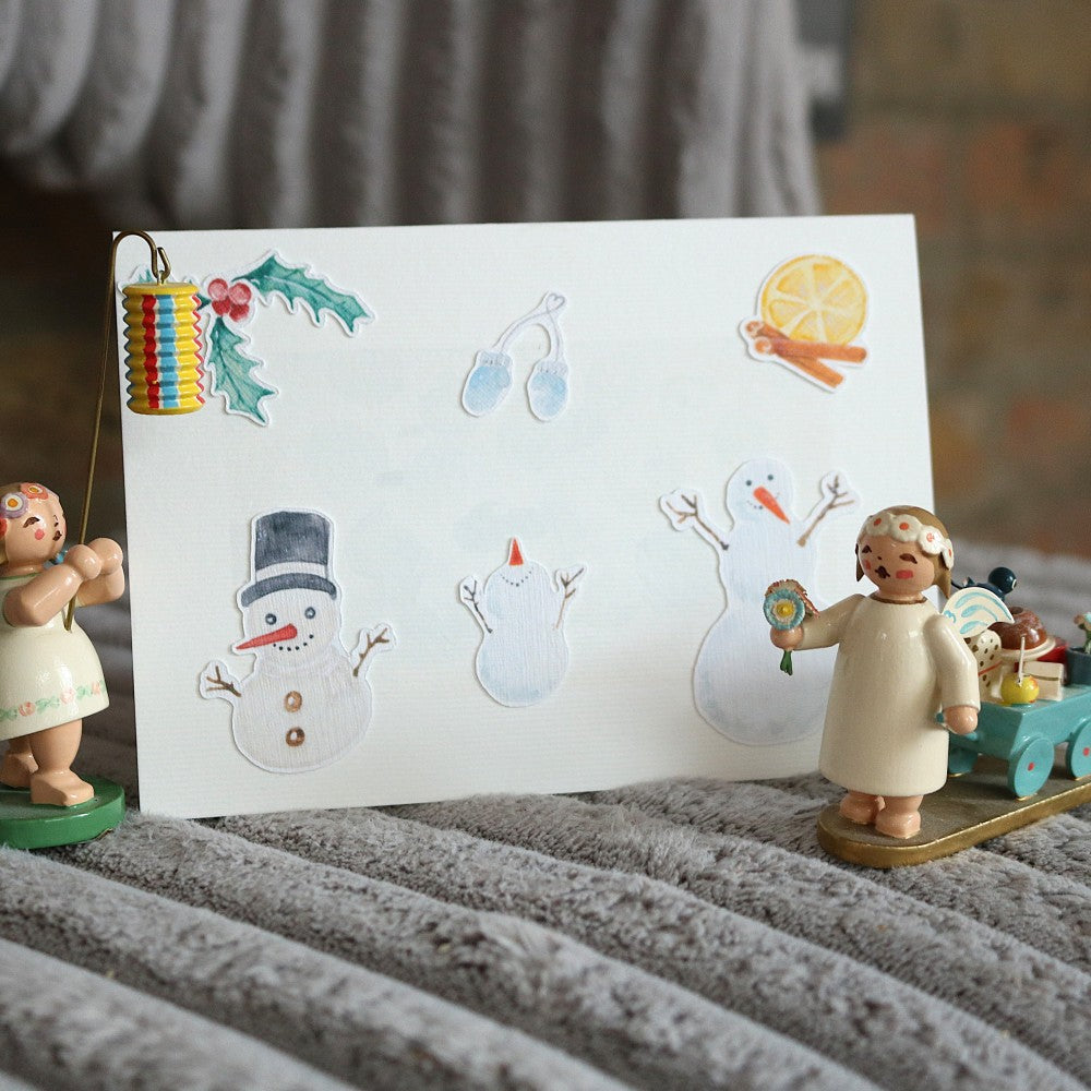 Klappkarte mit winterlichem Aquarell-Hintergrund, dekoriert mit ausgeschnittenen Motiven wie Schneemännern, Zitrone mit Zimtstange, Mistelzweig und Fäustlingen; im Vordergrund stehen zwei kleine Holzfiguren als Deko, die nicht zum Produkt gehören