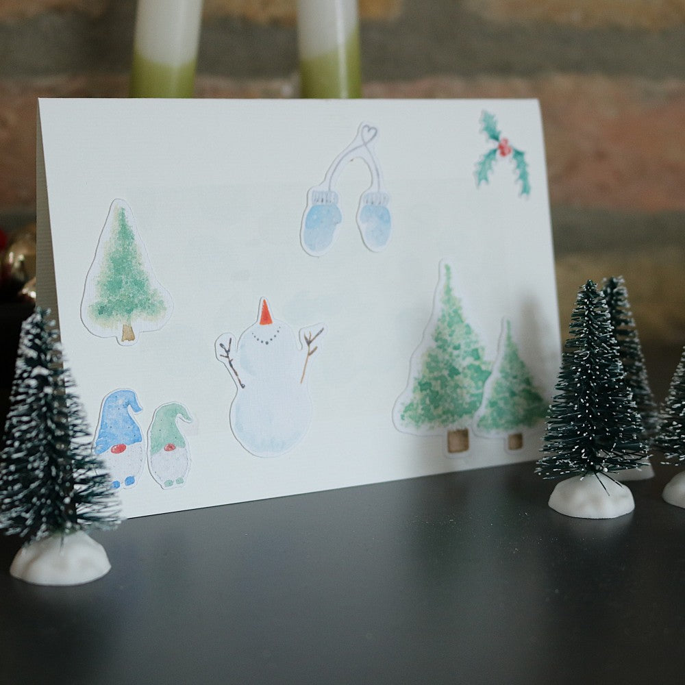 Klappkarte mit winterlichem Aquarell-Hintergrund, dekoriert mit ausgeschnittenen Weihnachtsmotiven wie Schneemann, Tannen, blauen Handschuhen, Mistelzweig und zwei kleinen Wichteln; die Karte steht zwischen Dekotannen vor einer rustikalen Steinwand