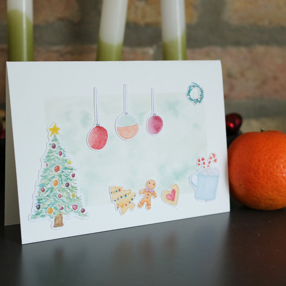 Klappkarte mit sanftem Aquarell-Hintergrund, verziert mit ausgeschnittenen Weihnachtsmotiven wie geschmücktem Tannenbaum, Christbaumkugeln, Lebkuchenmännchen, Plätzchen, Herz, kleinen Tannen und einer Tasse mit Zuckerstangen; daneben liegt eine Mandarine als Deko vor einer rustikalen Steinwand
