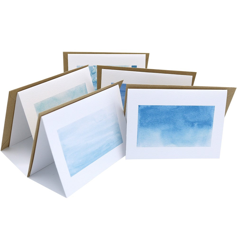 Aufgestellte Klappkarten aus dem Set Im Meer, jede mit einem anderen blauen Aquarellmotiv auf strukturiertem Papier. Dahinter liegen passende Kraftpapier-Umschläge. Die Karten stehen fächerförmig angeordnet und zeigen verschiedene ruhige Wasser- und Himmeltöne.