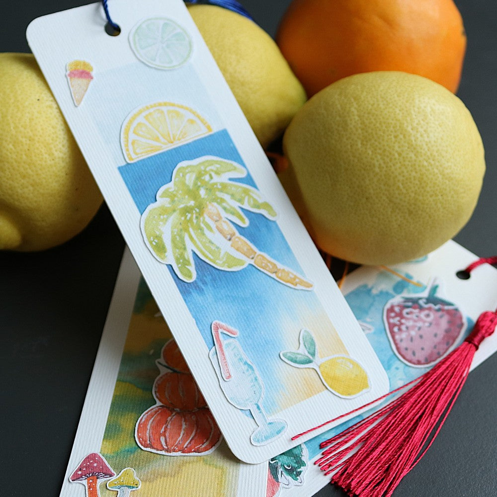 Drei Lesezeichen, eines aufgestellt mit Palmen und Obstscheiben, die anderen beiden darunter liegend; mit Quasten Lesezeichenbändchen